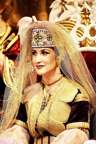 A woman in the traditional Moroccan-Jewish bridal dress — a gold-embroidered velvet caftan with an elaborate headdress of gold filigree and brocade — photographed at a traditional wedding ceremony in Morocco, showing the living tradition of Moroccan-Jewish celebratory dress